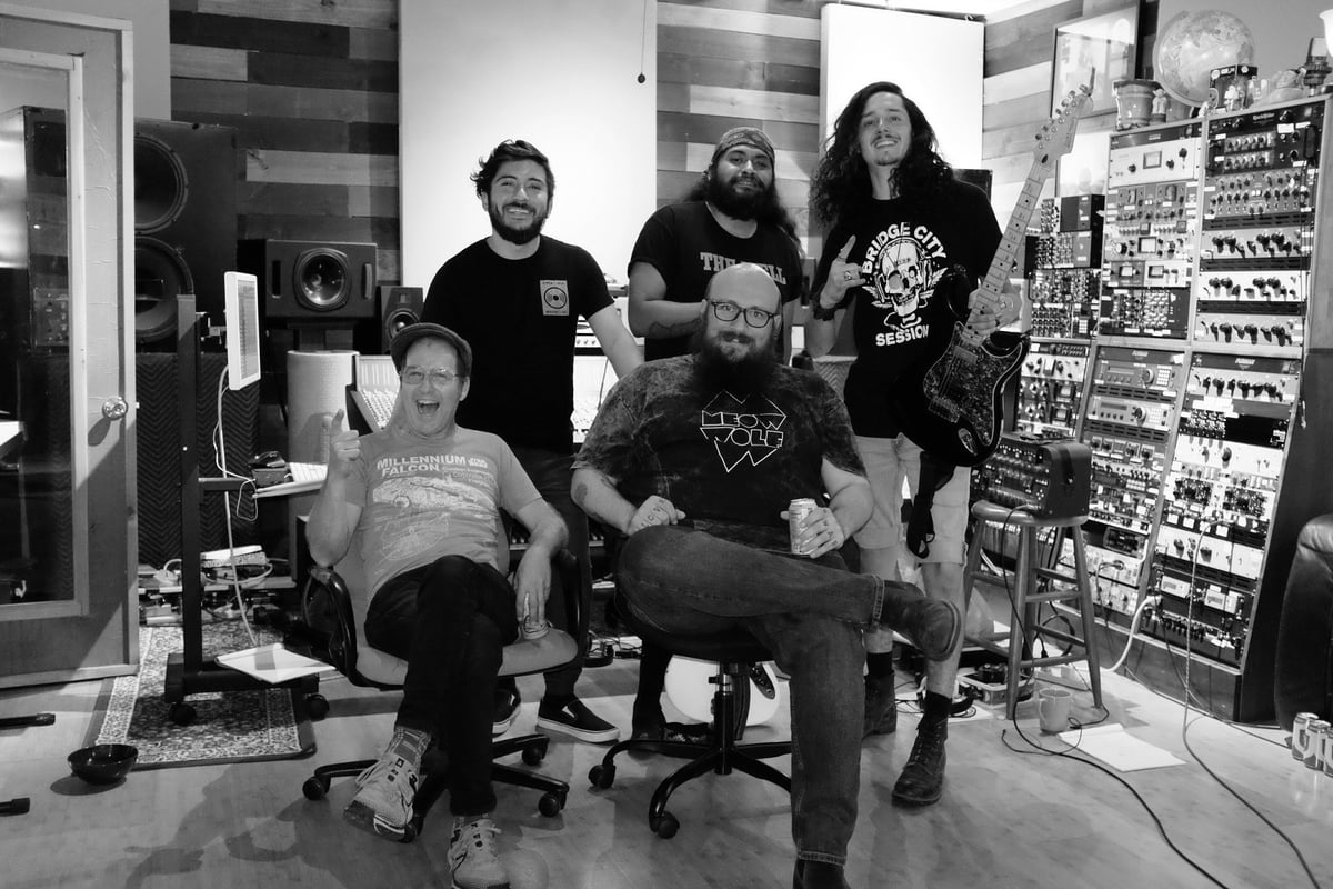 Five men posing in a recording studio, black and white photo. Band members in dark t-shirts with graphic designs stand and sit amid music equipment and posters on walls.