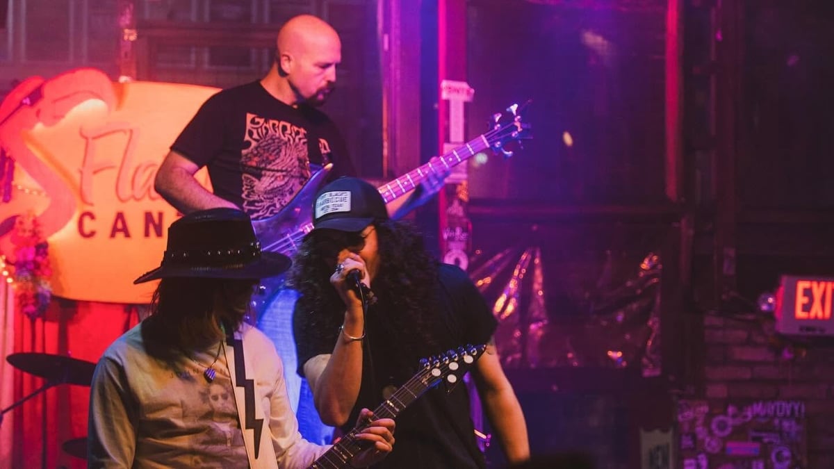 Bald musician playing electric guitar on stage under purple and pink neon lighting with audience members in foreground