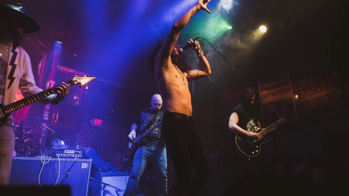 Live band performing on stage with blue and purple stage lighting, featuring singer at center microphone and guitarists on either side.