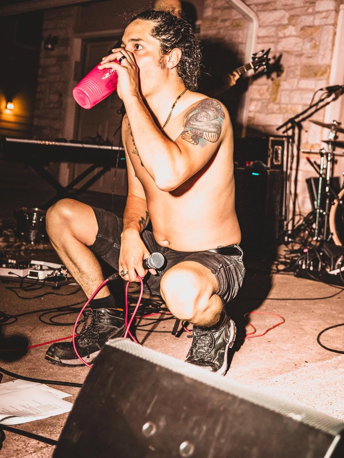 Shirtless male performer drinking from a pink cup while crouching on stage with band equipment and microphone stand visible
