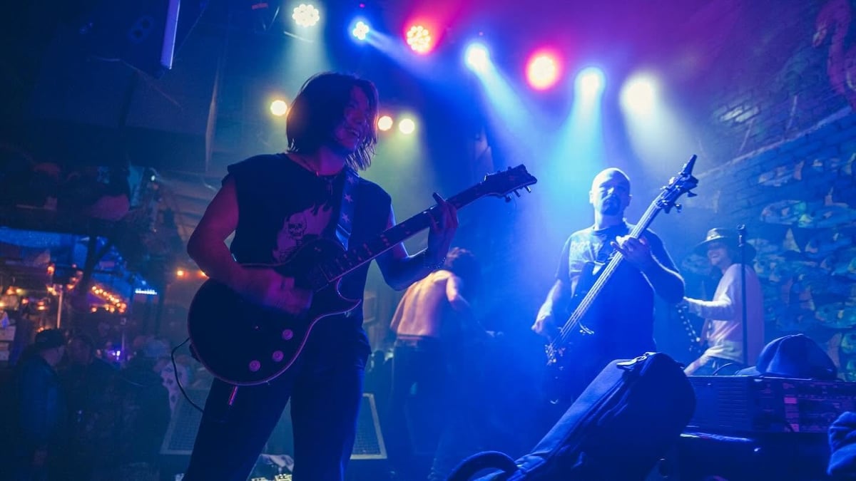 Live rock band performing on stage with guitarist in foreground, vibrant blue and purple stage lighting, and audience members visible below.