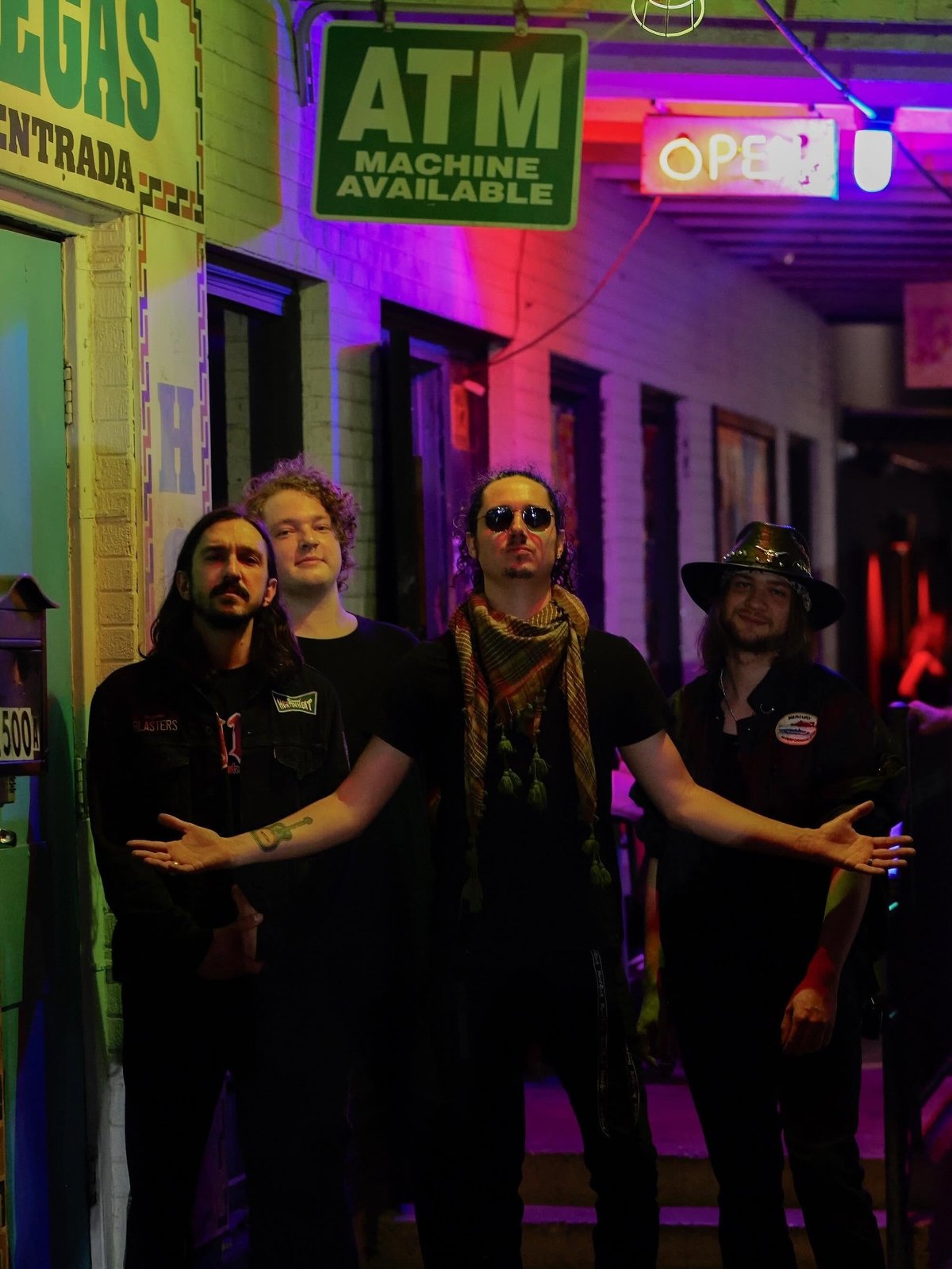 Four band members standing on a neon-lit street with ATM and open signs, wearing black clothes and sunglasses in a vibrant purple and pink atmosphere.