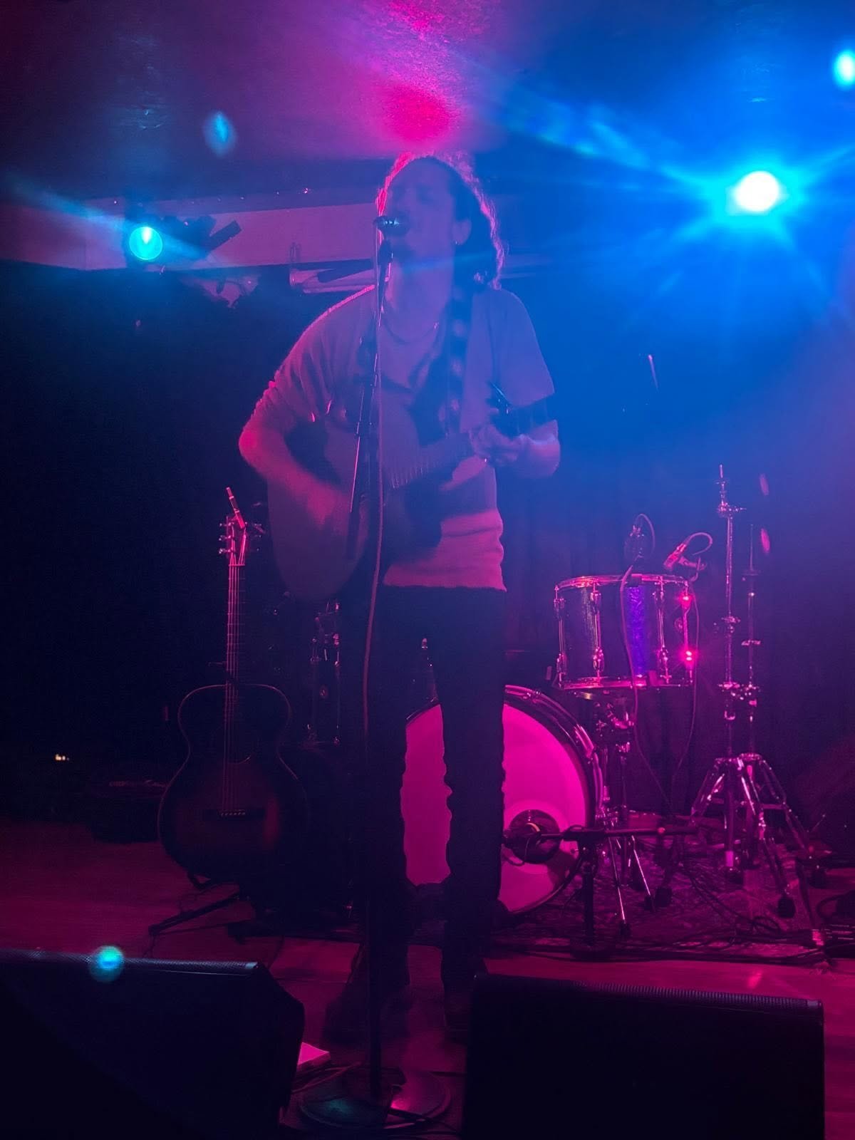 Male musician performing on stage with acoustic guitar, illuminated by blue and pink stage lights with drum kit visible in background