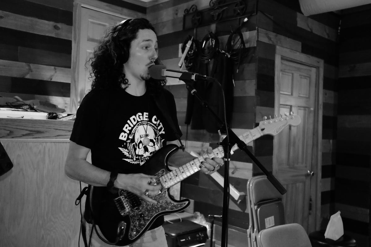 Man with long dark hair playing electric guitar in a home studio, wearing a black graphic t-shirt, black and white photograph