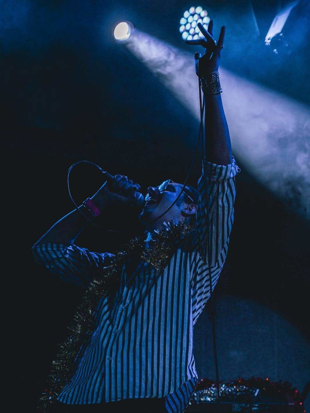 Performer on stage in blue striped outfit with arm raised toward stage lights during concert performance