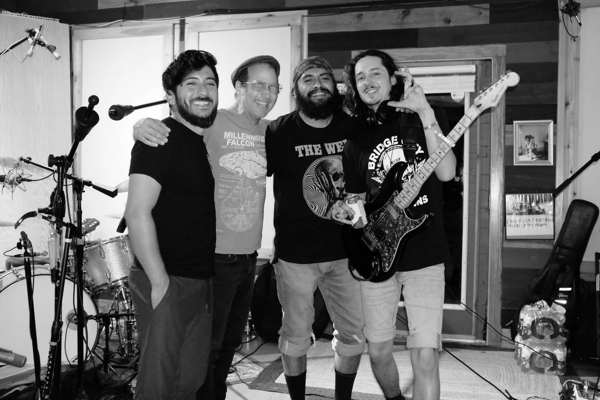 Four band members posing together in a studio with drums, guitar, and equipment visible in the background.