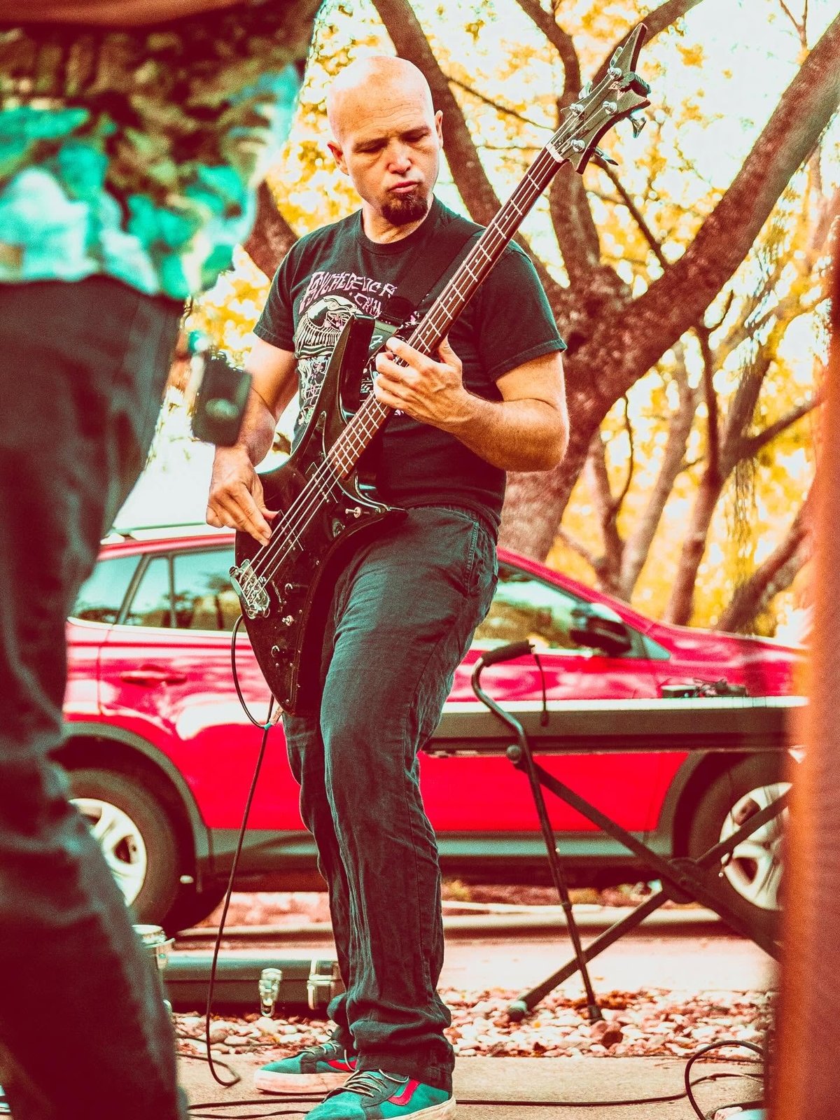 Bald musician playing bass guitar outdoors, wearing dark band t-shirt, with red vintage car and autumn trees in background