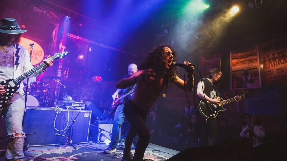 Live band performing on stage with colorful blue, purple, and yellow stage lighting, featuring vocalist, bassist, guitarist, and drummer
