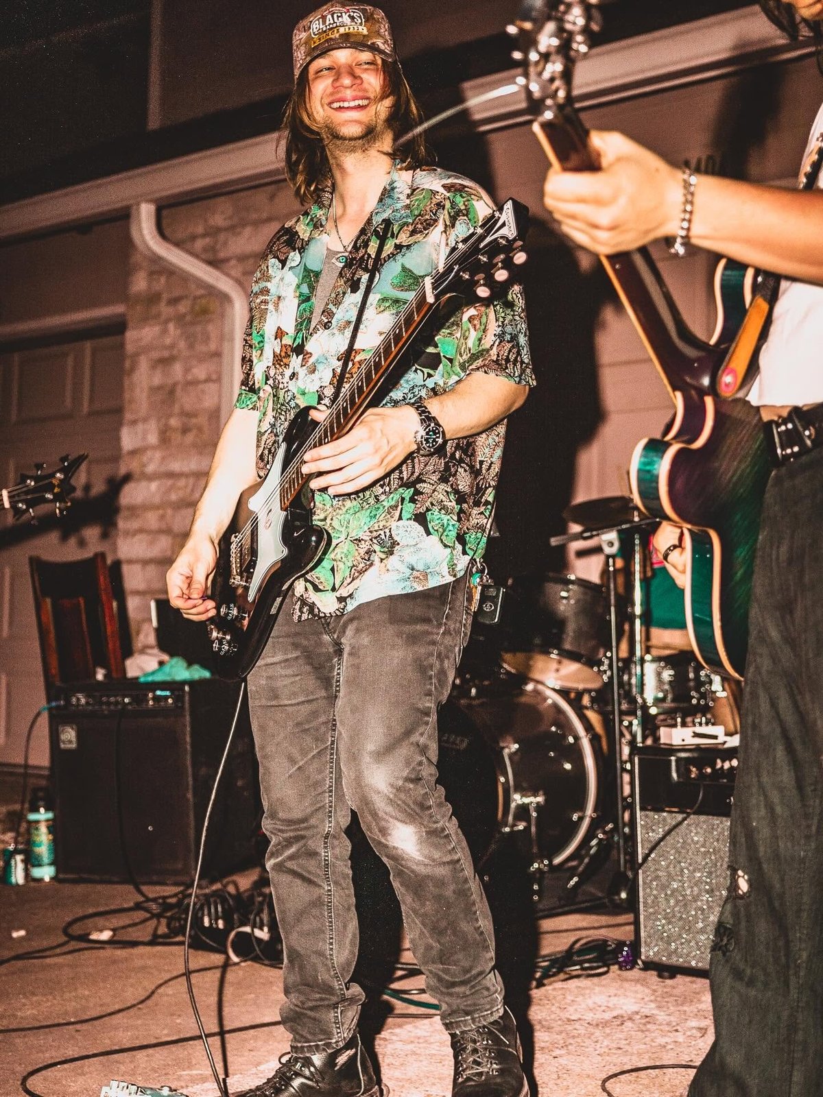 Male musician performing with electric guitar on stage, wearing colorful floral shirt and headband, smiling at audience