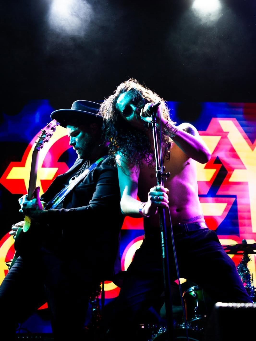 Musicians performing on stage with vibrant colorful neon lighting and geometric designs in the background