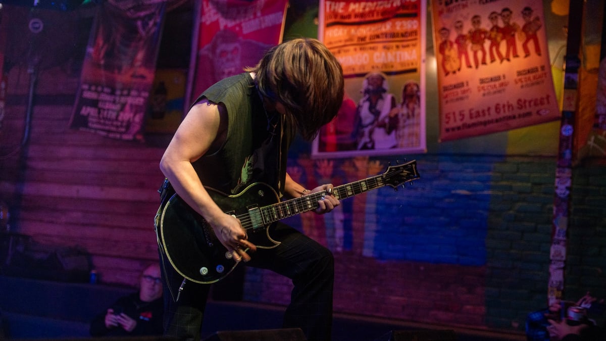 Guitarist performing on stage under purple and blue lighting, playing a black electric guitar with vintage posters in the background