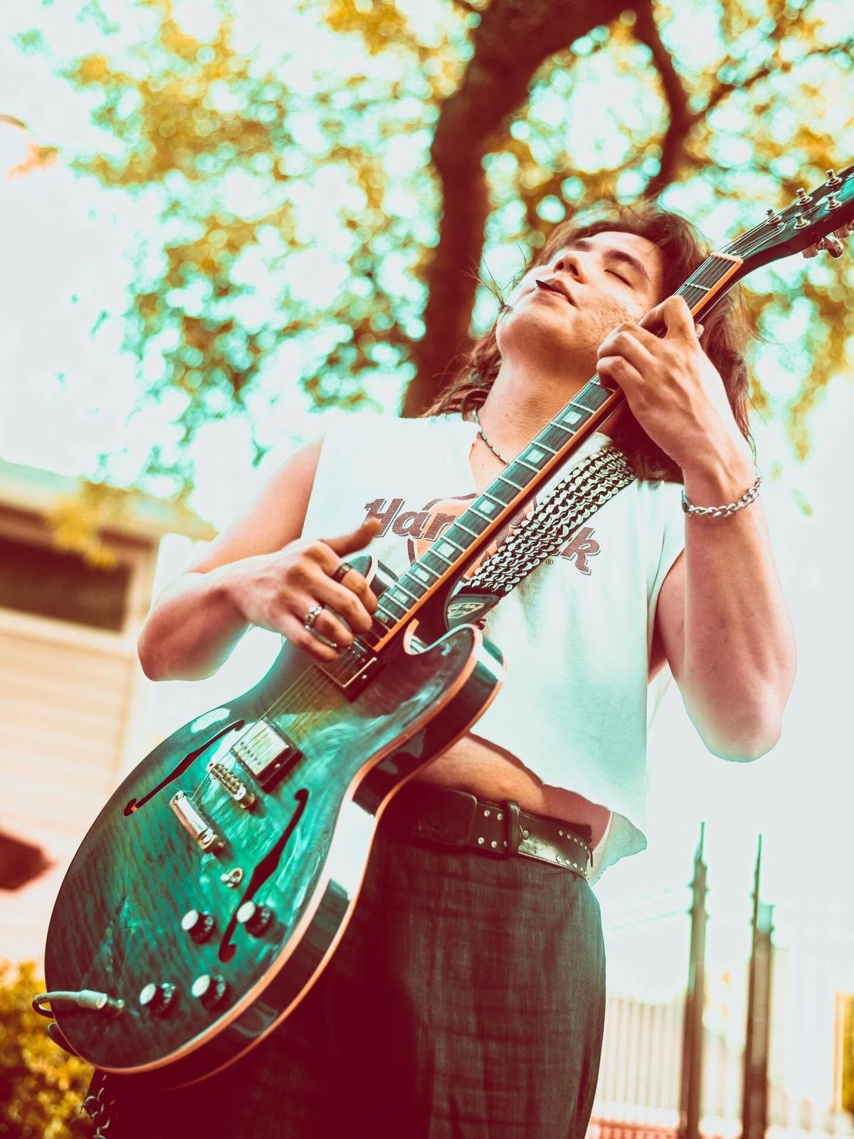 Young musician playing electric guitar outdoors with tree canopy and urban background, looking upward