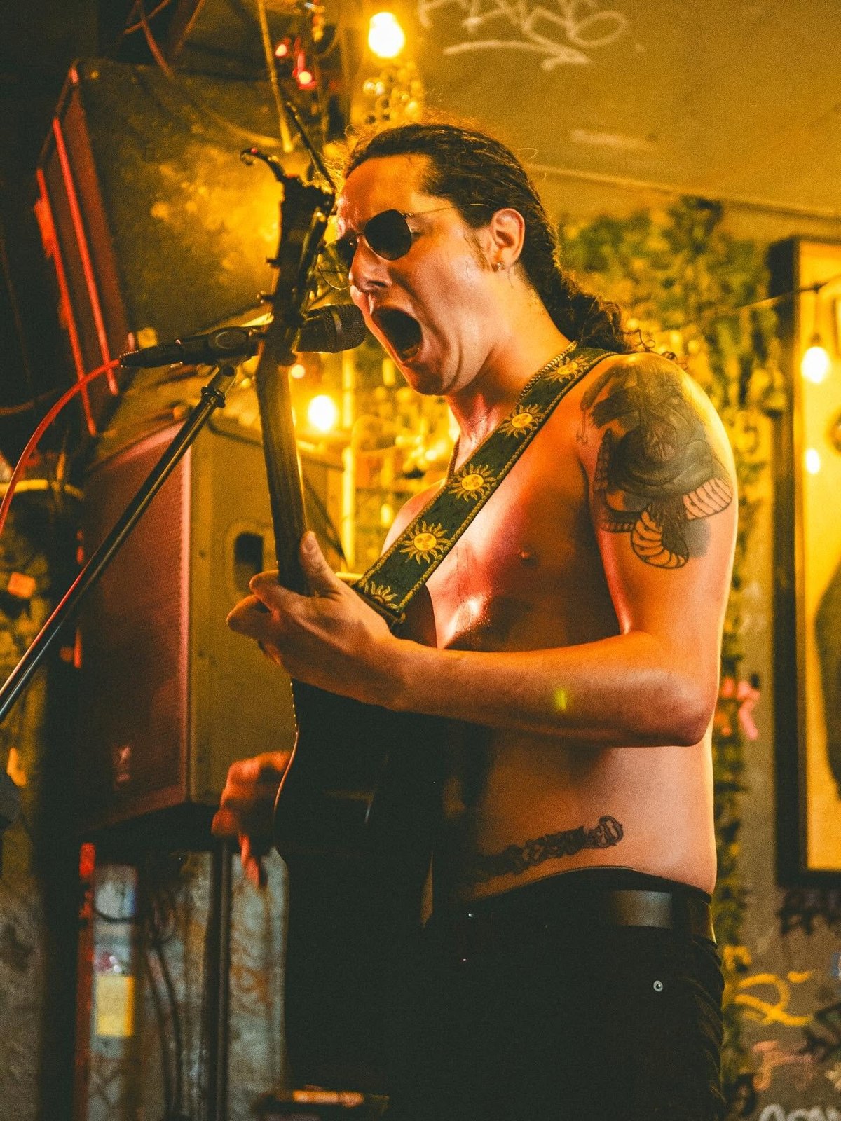 Male musician performing on stage, singing into microphone with sunglasses and tattoos, warm stage lighting with yellow tones