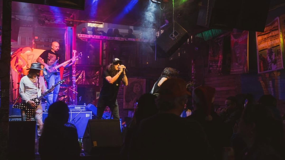 Live band performing on stage in a dark venue with neon pink and blue lighting, audience members silhouetted in foreground
