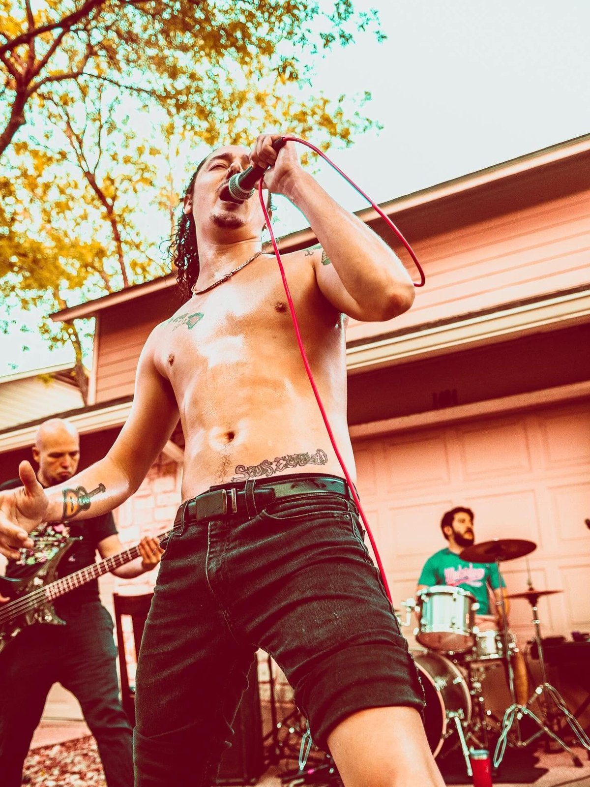 Shirtless male vocalist singing into microphone with band performing behind him outdoors near a house