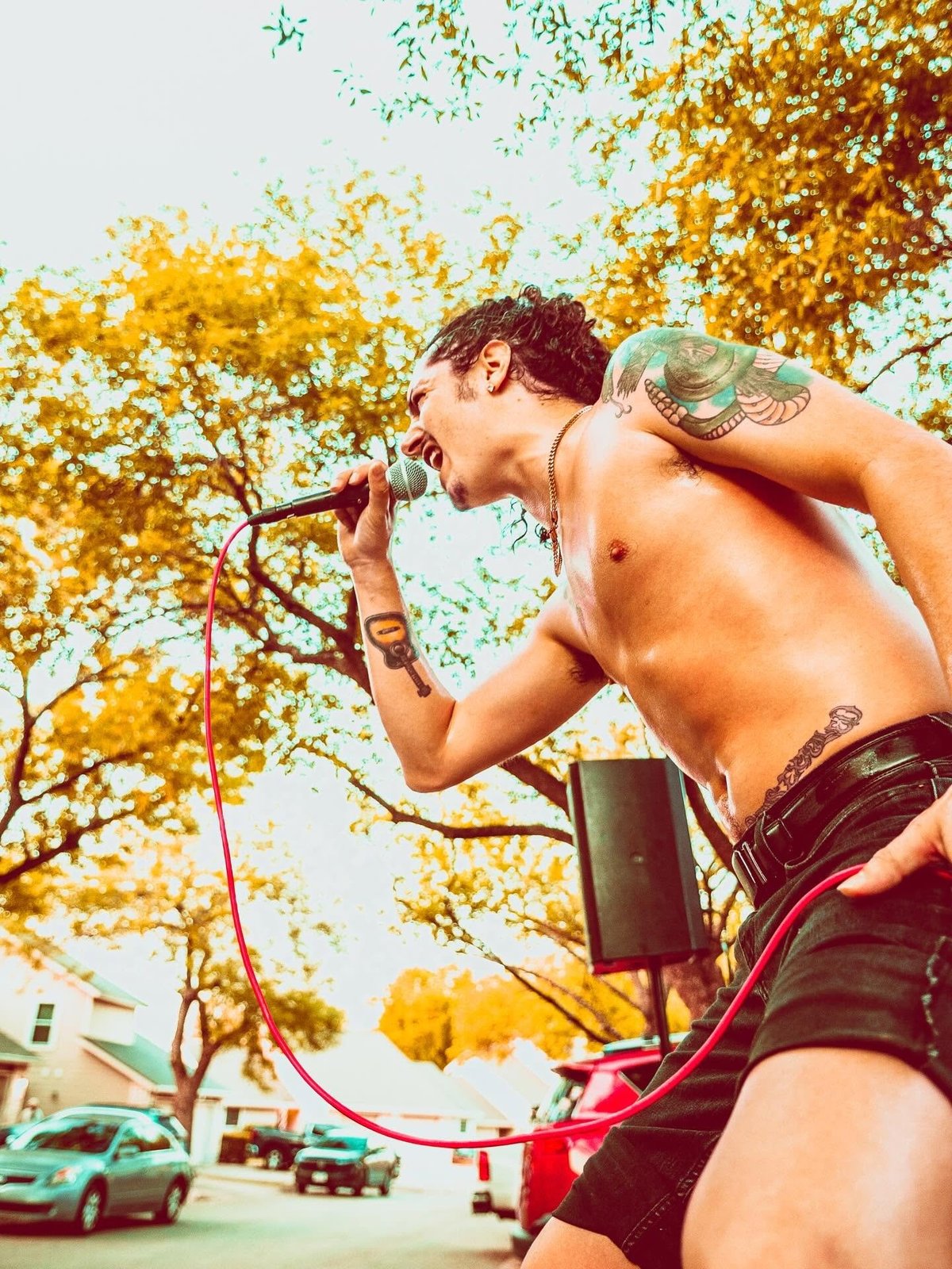 Shirtless man singing into a microphone outdoors with fall foliage and trees in the background