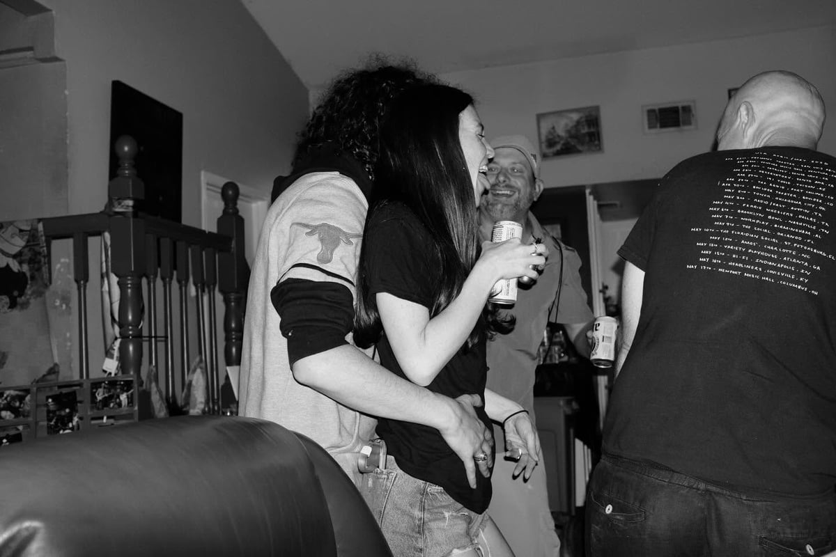 Black and white photo of a woman with long dark hair holding a microphone, surrounded by people at an indoor event or gathering.
