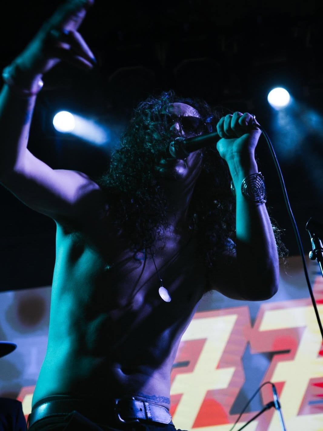 Male performer singing into microphone on stage with blue and purple stage lighting and colorful backdrop