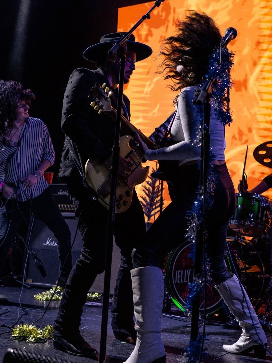 Musicians performing on stage with guitars, bright orange backdrop lighting, band members in dark clothing with sparkly embellishments