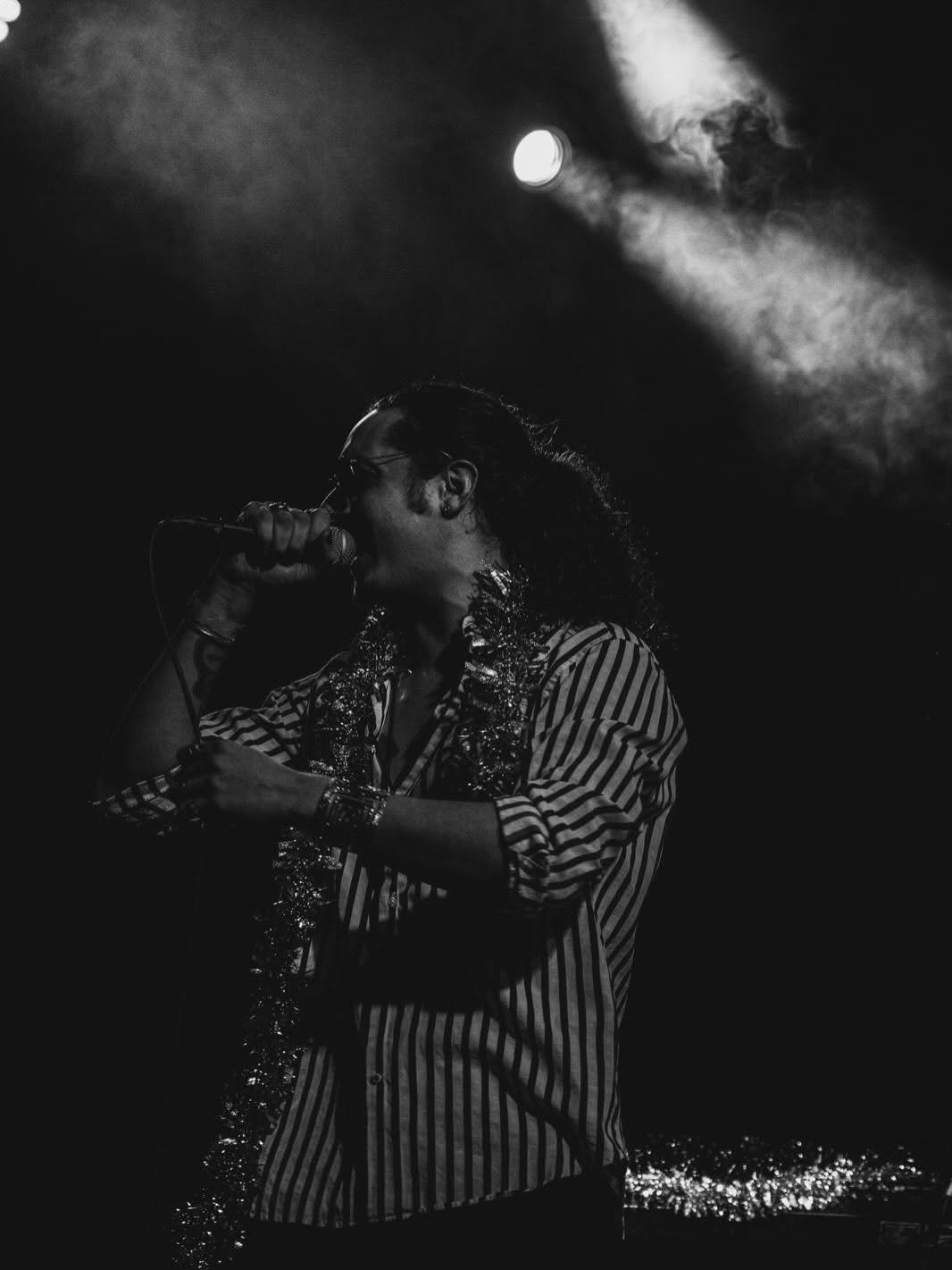 Black and white image of a performer singing on stage with dramatic spotlight lighting and layered clothing