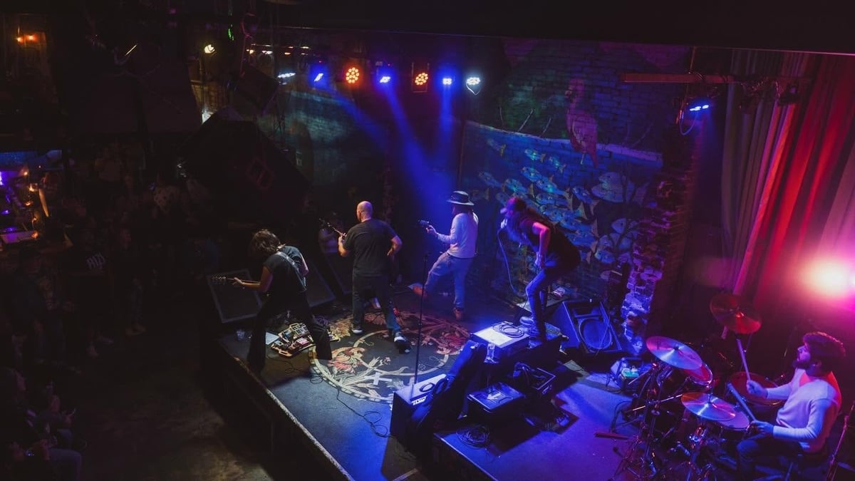 Live band performing on stage in a dark venue with blue and pink stage lighting and an audience watching