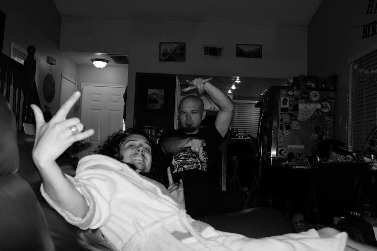 Black and white photo of two men in a cluttered room, one lying down making peace signs while another stands behind him.