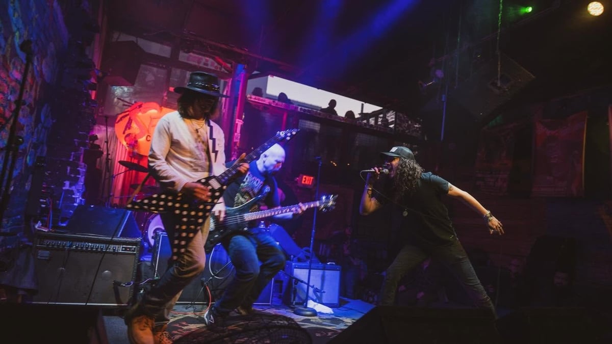 Live rock band performing on stage with blue and red stage lighting, guitarist in patterned pants and audience member in foreground