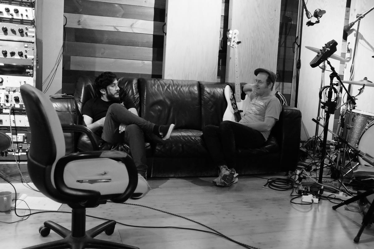 Two men sitting on a black couch in a recording studio with professional equipment and microphones visible