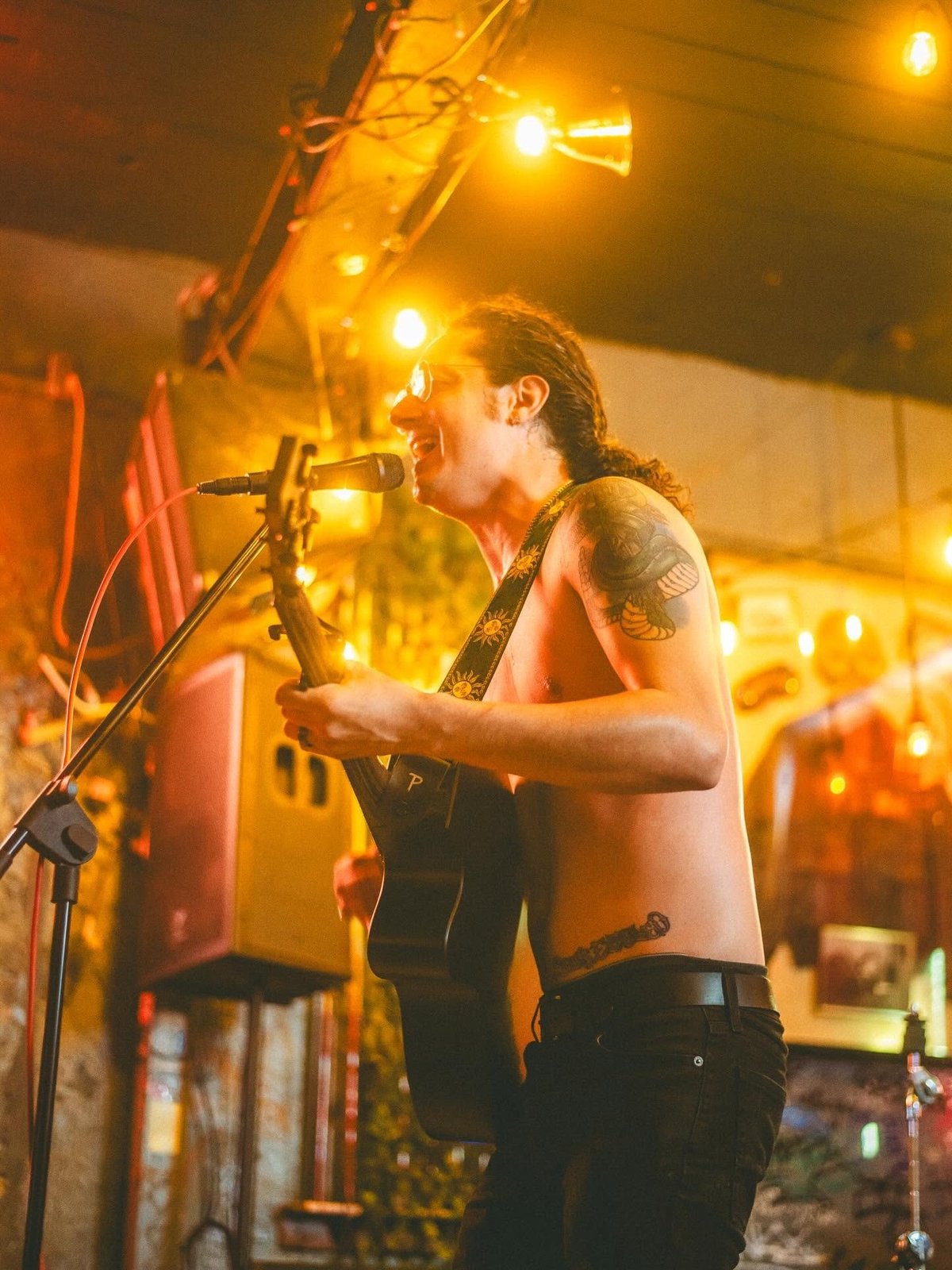 Shirtless male musician performing at a microphone on stage with warm yellow stage lighting overhead