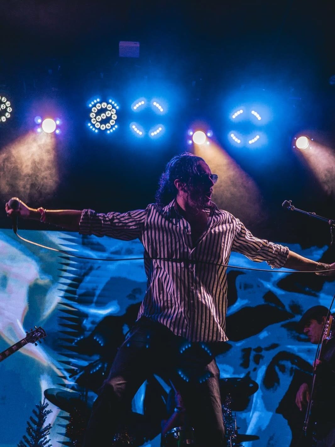 A performer on stage wearing a striped shirt with arms outstretched, illuminated by blue and purple stage lighting and geometric light shapes