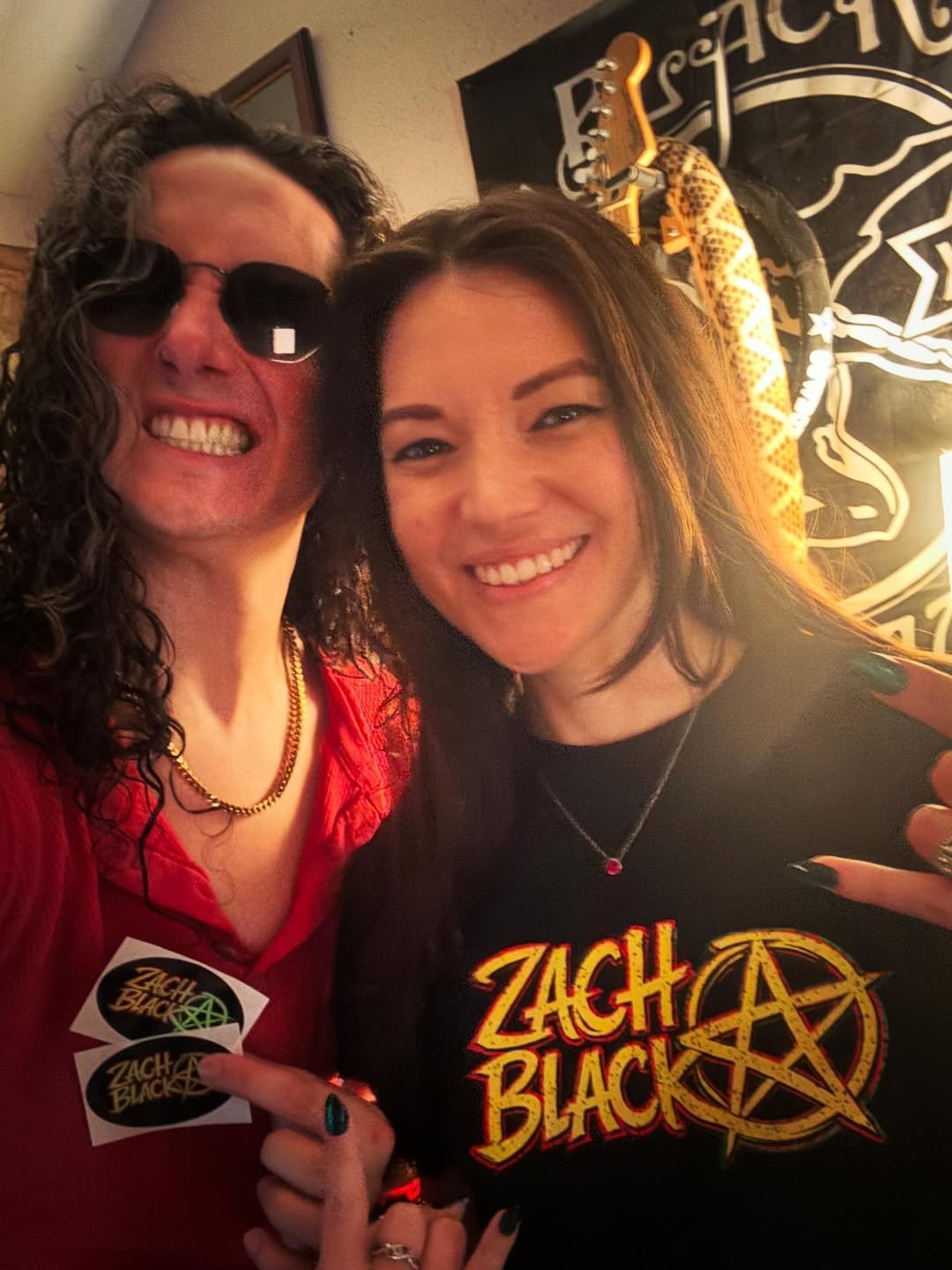 Two people smiling at camera, one wearing sunglasses and red shirt, the other in black Zach Black band shirt, holding stickers indoors