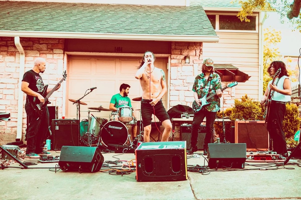 A band performs outdoors in front of a brick house, with musicians playing guitar, drums, and vocals on a driveway stage.