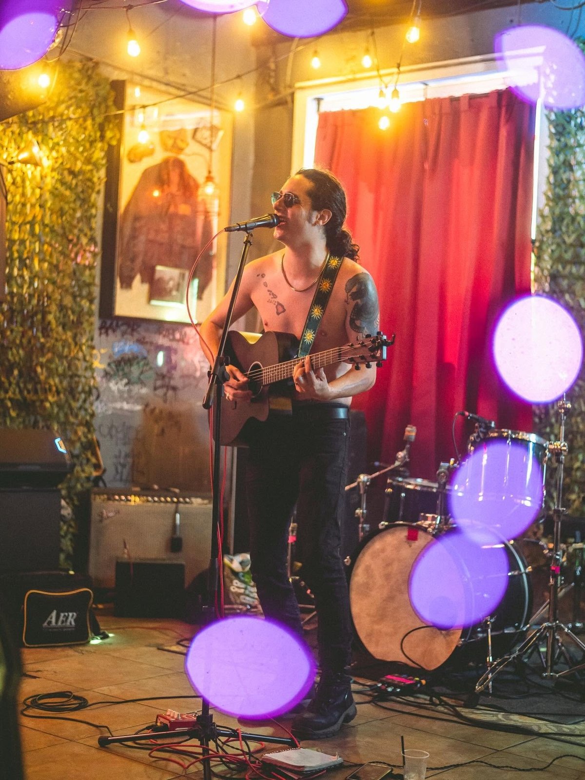 Female musician performing on stage with guitar and microphone, surrounded by purple and golden stage lights and drum kit.