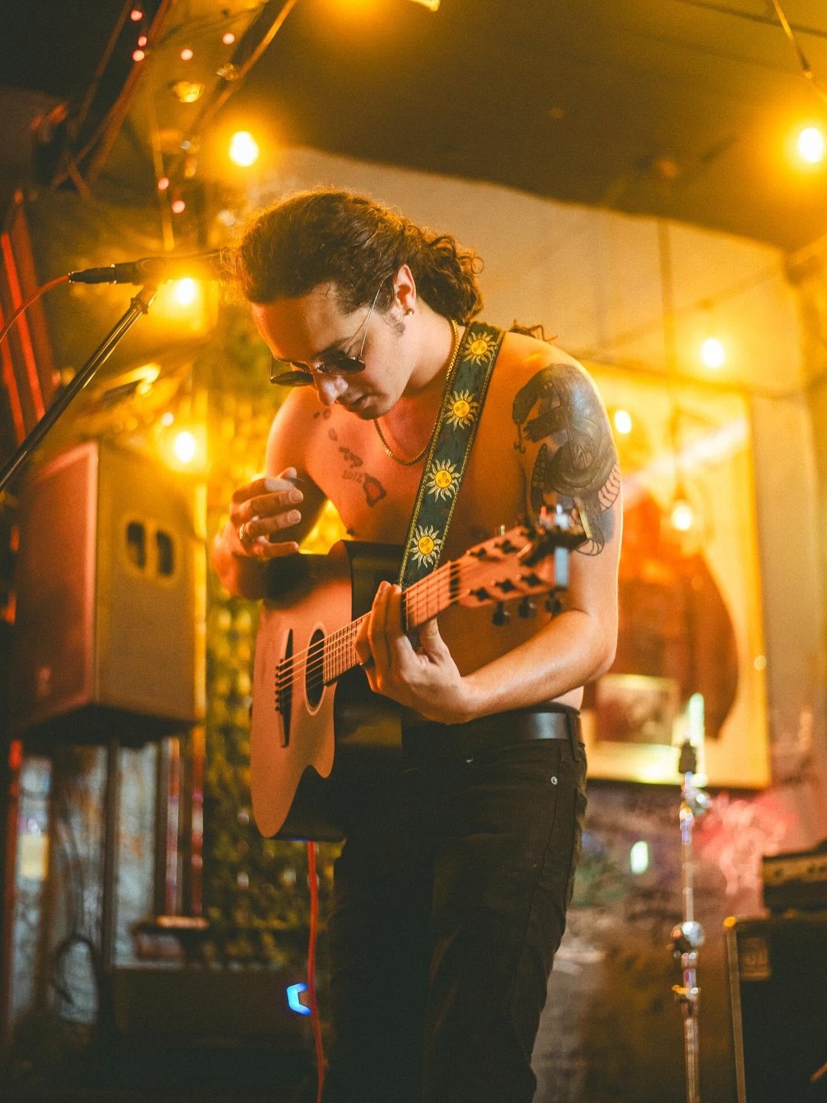 Musician wearing sunglasses and a patterned guitar strap performing on stage with warm stage lighting overhead