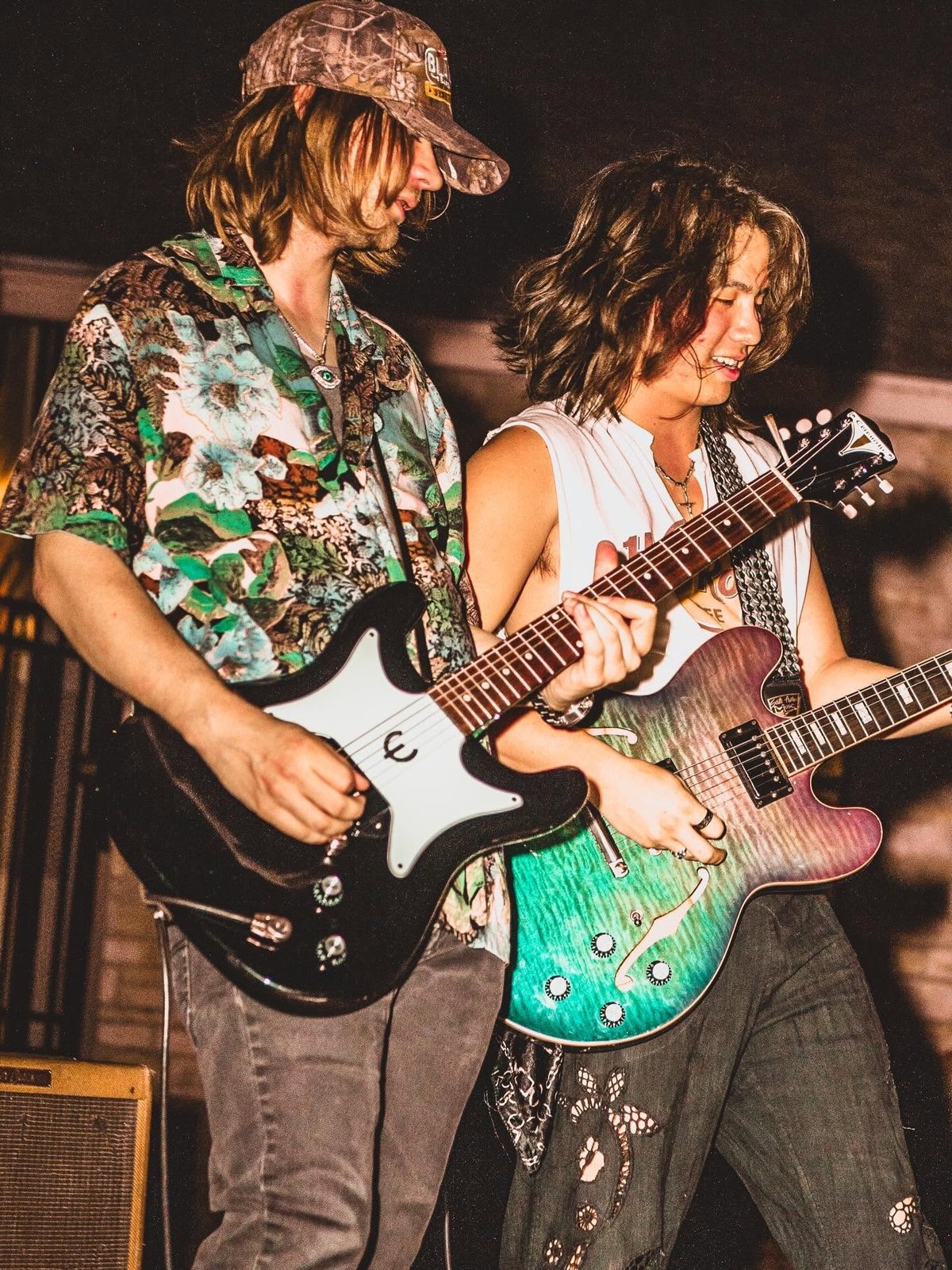 Two musicians performing on stage, one playing a black and white electric guitar wearing a patterned shirt and cap, the other playing a green guitar in white tank top.