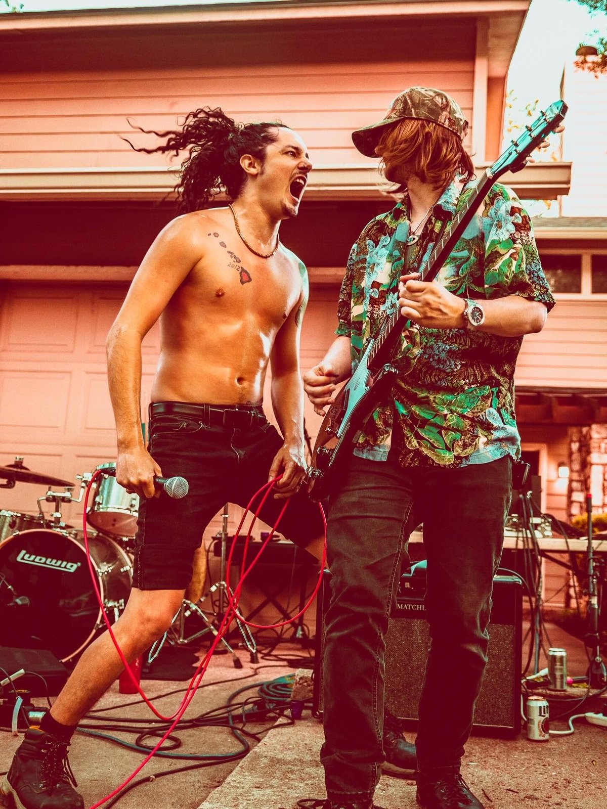 Two musicians performing in a garage with instruments, one shirtless with dreadlocks and one wearing a patterned shirt holding a guitar