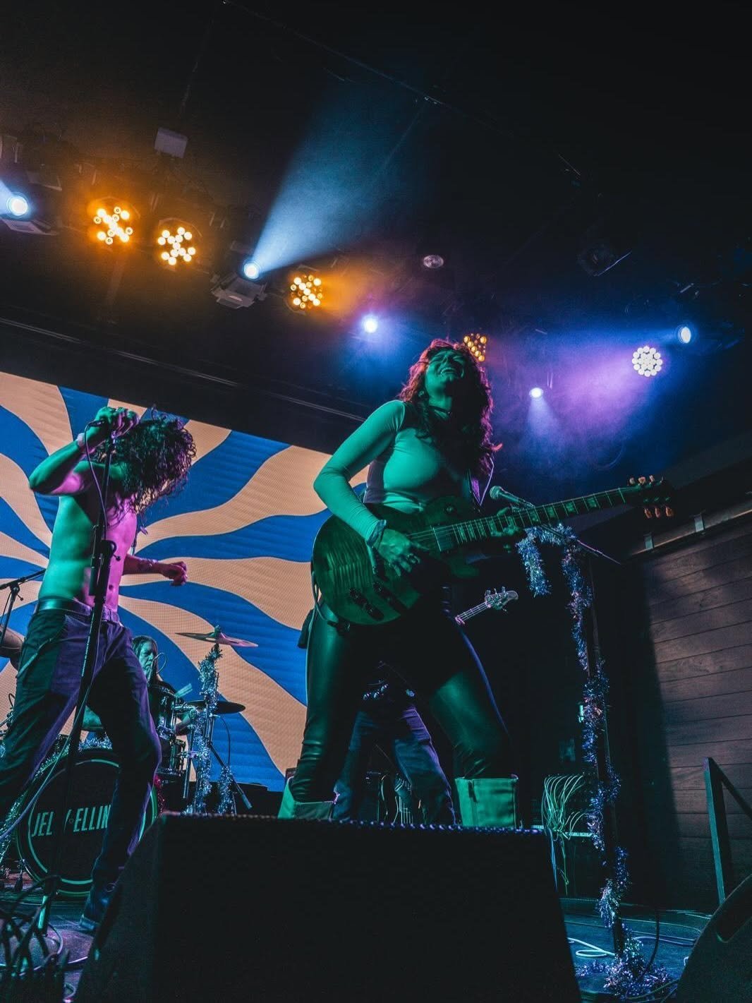 Female guitarist performs on stage with colorful stage lighting, green and blue spotlights illuminating the band during a concert performance.