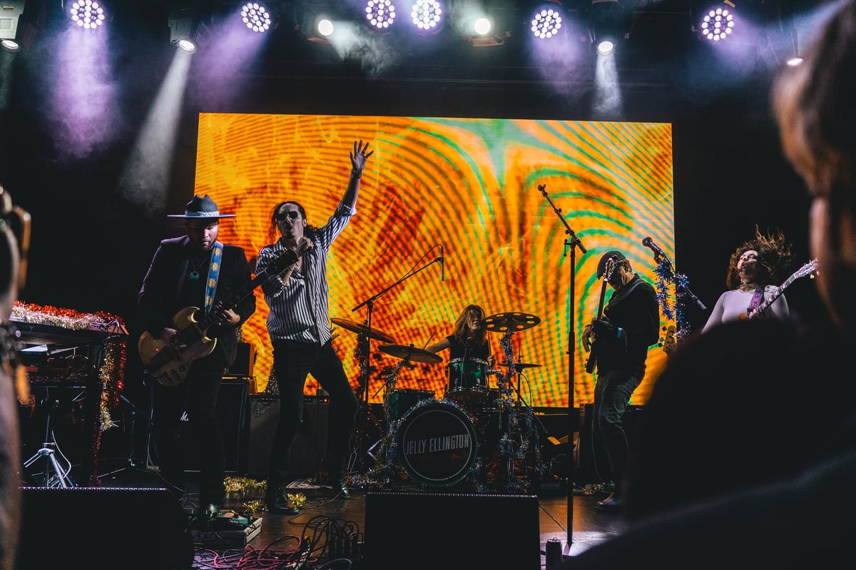 Band performing on stage with colorful yellow and orange visual display behind them, purple stage lighting, and audience members visible in foreground
