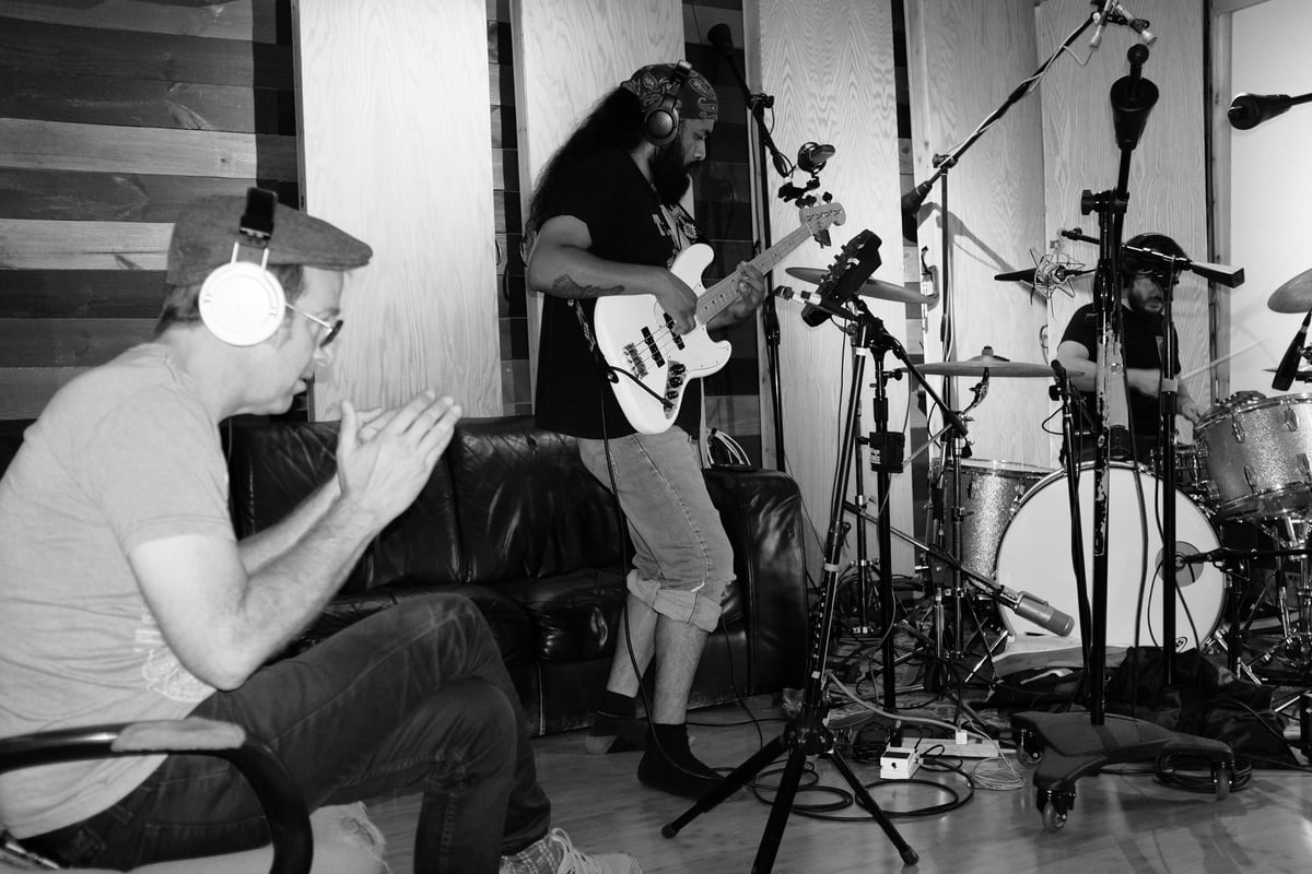 Black and white photo of two musicians performing in a studio. One plays guitar while the other sings into a microphone, surrounded by drums and recording equipment.